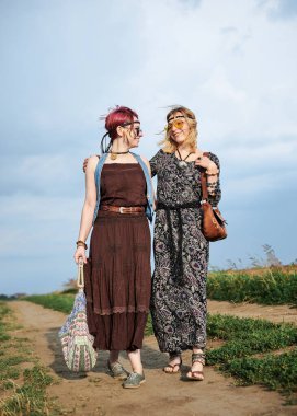 İki hippi kadın, boho tarzı kıyafetler giyiyorlar, yeşil alanda toprak yolda yürüyorlar, eğleniyorlar. Kırsalda birlikte seyahat eden bayan arkadaşlar. Eko turizm konsepti. Yaz tatili boş zaman.