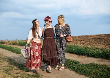 Üç hippi kadın, boho tarzı kıyafetler giyiyorlar, yeşil alanda toprak yolda yürüyorlar, eğleniyorlar. Kırsalda birlikte seyahat eden bayan arkadaşlar. Eko turizm konsepti. Yaz tatili boş zaman.