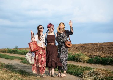 Üç hippi kadın, boho tarzı kıyafetler giyiyorlar, yeşil alanda toprak yolda yürüyorlar, eğleniyorlar. Kırsalda birlikte seyahat eden bayan arkadaşlar. Eko turizm konsepti. Yaz tatili boş zaman.