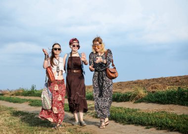 Üç hippi kadın, boho tarzı kıyafetler giyiyorlar, yeşil alanda toprak yolda yürüyorlar, eğleniyorlar. Kırsalda birlikte seyahat eden bayan arkadaşlar. Eko turizm konsepti. Yaz tatili boş zaman.