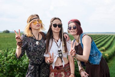 Üç hippi kadın, boho tarzı kıyafetler giyiyorlar, yeşil frenk üzümü tarlasında barış işareti gösteriyorlar, eğleniyorlar. Kırsalda birlikte seyahat eden bayan arkadaşlar. Eko turizm konsepti. Yaz tatili boş zaman