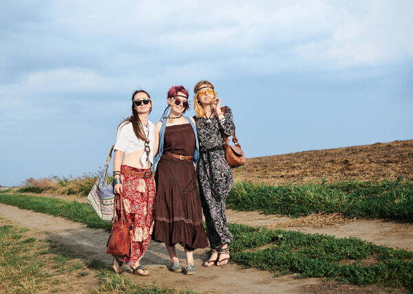 Three hippie women, wearing boho style clothes, walking on dirt road on green field, having fun. Female friends, traveling together in countryside. Eco tourism concept. Summer leisure free time.