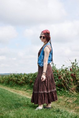 Kısa kızıl saçlı, boho tarzı kıyafetler ve güneş gözlüğü takan, yeşil frenk üzümü tarlasında duran, resim için poz veren genç hippi kadın. Doğal arka planda bir kadın portresi. Çevre turizmi kavramı.