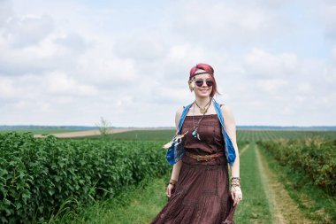 Kısa kızıl saçlı, boho tarzı kıyafetler ve güneş gözlüğü takan, yeşil frenk üzümü tarlasında dans eden, resim için poz veren genç hippi kadın. Doğal arka planda bir kadın portresi. Çevre turizmi kavramı.