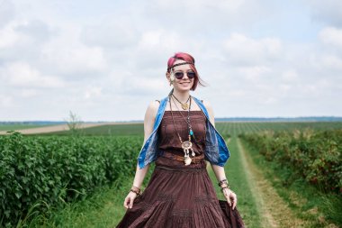 Kısa kızıl saçlı, boho tarzı kıyafetler ve güneş gözlüğü takan, yeşil frenk üzümü tarlasında dans eden, resim için poz veren genç hippi kadın. Doğal arka planda bir kadın portresi. Çevre turizmi kavramı.
