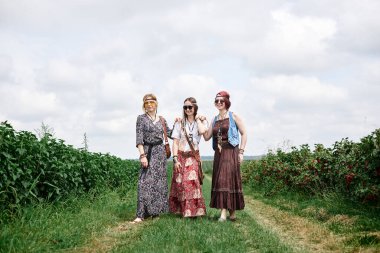 Üç genç hippi kadın, boho tarzı kıyafetler giyiyorlar, yazın yeşil frenk üzümü tarlasında yürüyorlar. Eko turizm konsepti. Kırsal kesimde seyahat eden arkadaşlar.