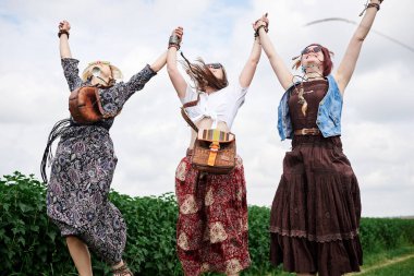 Üç genç hippi kadın, boho tarzı kıyafetler giyiyorlar, atlıyorlar, yazın yeşil frenk üzümü tarlasında el ele tutuşuyorlar. Eko turizm konsepti. Kırsal kesimde seyahat eden arkadaşlar.