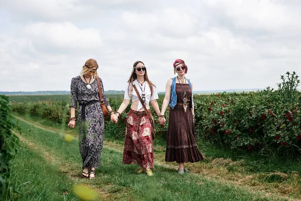Üç genç hippi kadın, boho tarzı kıyafetler giyiyorlar, el ele tutuşuyorlar, yazın yeşil frenk üzümü tarlasında dans ediyorlar. Eko turizm konsepti. Kırsal kesimde seyahat eden arkadaşlar.