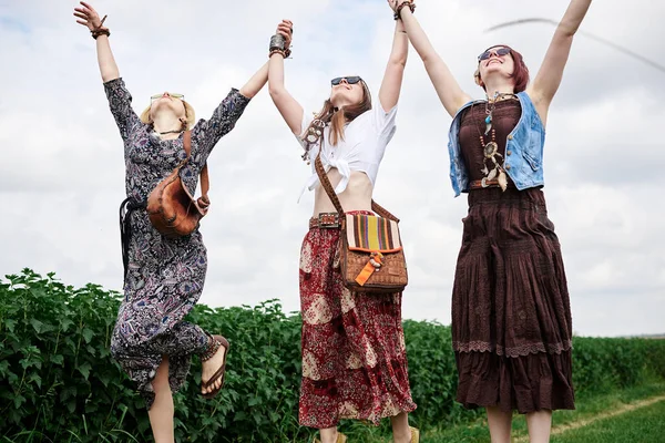 Üç genç hippi kadın, boho tarzı kıyafetler giyiyorlar, atlıyorlar, yazın yeşil frenk üzümü tarlasında el ele tutuşuyorlar. Eko turizm konsepti. Kırsal kesimde seyahat eden arkadaşlar.