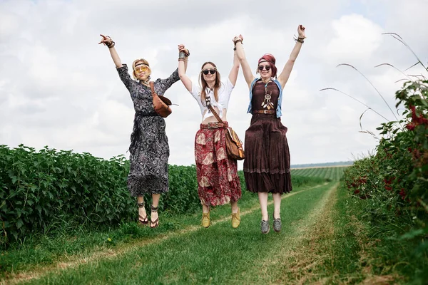 Üç genç hippi kadın, boho tarzı kıyafetler giyiyorlar, atlıyorlar, yazın yeşil frenk üzümü tarlasında el ele tutuşuyorlar. Eko turizm konsepti. Kırsal kesimde seyahat eden arkadaşlar.