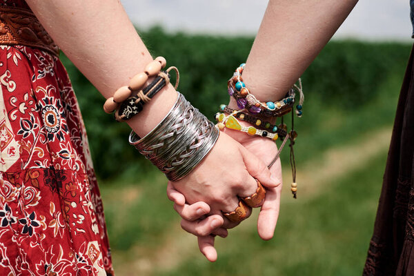 Close-up picture of hands with colorful bracelets and various rings, holding each other. Hippie women, wearing boho style clothes, standing on green field. Female friendship concept.