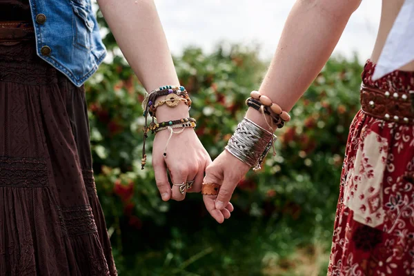 Renkli bileziklerle ve çeşitli yüzüklerle birbirine sarılmış ellerin yakın çekimi. Hippi kadınlar, boho tarzı kıyafetler giyiyorlar, yeşil alanda duruyorlar. Kadın arkadaşlığı kavramı.