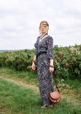 Genç sarışın hippi kadın, gri boho tarzı elbise ve sarı güneş gözlüğü takıyor, kahverengi deri çanta tutuyor, yeşil alanda duruyor, güneşli yaz gününde fotoğraf için poz veriyor. İç özgürlük kavramı.