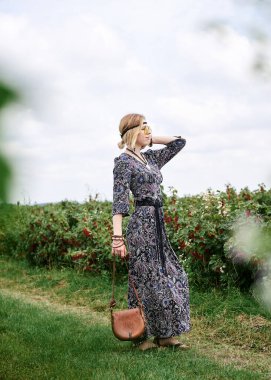 Genç sarışın hippi kadın, gri boho tarzı elbise ve sarı güneş gözlüğü takıyor, kahverengi deri çanta tutuyor, yeşil alanda duruyor, güneşli yaz gününde fotoğraf için poz veriyor. İç özgürlük kavramı.