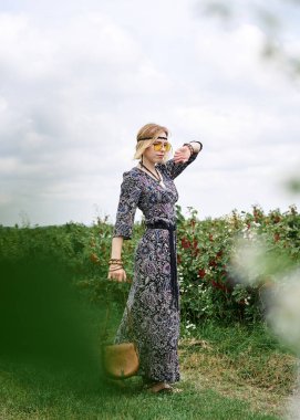 Genç sarışın hippi kadın, gri boho tarzı elbise ve sarı güneş gözlüğü takıyor, kahverengi deri çanta tutuyor, yeşil alanda duruyor, güneşli yaz gününde fotoğraf için poz veriyor. İç özgürlük kavramı.