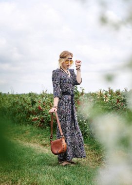 Genç sarışın hippi kadın, gri boho tarzı elbise ve sarı güneş gözlüğü takıyor, kahverengi deri çanta tutuyor, yeşil alanda duruyor, güneşli yaz gününde fotoğraf için poz veriyor. İç özgürlük kavramı.