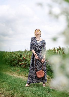 Genç sarışın hippi kadın, gri boho tarzı elbise ve sarı güneş gözlüğü takıyor, kahverengi deri çanta tutuyor, yeşil alanda duruyor, güneşli yaz gününde fotoğraf için poz veriyor. İç özgürlük kavramı.
