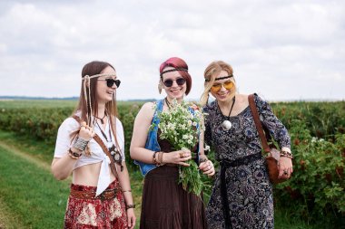 Üç genç hippi kadın, boho tarzı kıyafetler giyiyorlar, yeşil frenk üzümü tarlasında duruyorlar, yazın papatya buketi tutuyorlar. Eko turizm konsepti. Kırsal kesimde seyahat eden arkadaşlar.