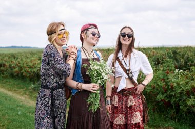 Üç genç hippi kadın, boho tarzı kıyafetler giyiyorlar, yeşil frenk üzümü tarlasında duruyorlar, yazın papatya buketi tutuyorlar. Eko turizm konsepti. Kırsal kesimde seyahat eden arkadaşlar.