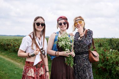 Üç genç hippi kadın, boho tarzı kıyafetler giyiyorlar, yeşil frenk üzümü tarlasında duruyorlar, yazın papatya buketi tutuyorlar. Eko turizm konsepti. Kırsal kesimde seyahat eden arkadaşlar.