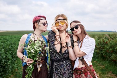 Üç genç hippi kadın, boho tarzı kıyafetler giyiyorlar, yeşil frenk üzümü tarlasında duruyorlar, yazın papatya buketi tutuyorlar. Eko turizm konsepti. Kırsal kesimde seyahat eden arkadaşlar.