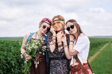 Üç genç hippi kadın, boho tarzı kıyafetler giyiyorlar, yeşil frenk üzümü tarlasında duruyorlar, yazın papatya buketi tutuyorlar. Eko turizm konsepti. Kırsal kesimde seyahat eden arkadaşlar.