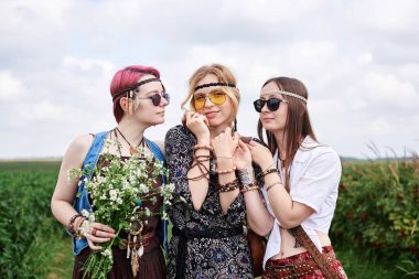 Üç genç hippi kadın, boho tarzı kıyafetler giyiyorlar, yeşil frenk üzümü tarlasında duruyorlar, yazın papatya buketi tutuyorlar. Eko turizm konsepti. Kırsal kesimde seyahat eden arkadaşlar.