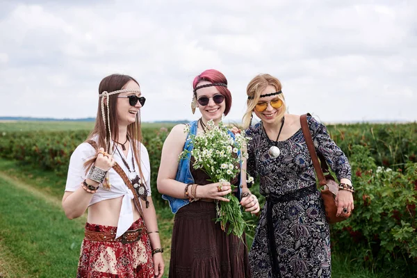 Üç genç hippi kadın, boho tarzı kıyafetler giyiyorlar, yeşil frenk üzümü tarlasında duruyorlar, yazın papatya buketi tutuyorlar. Eko turizm konsepti. Kırsal kesimde seyahat eden arkadaşlar.