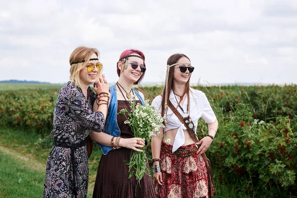 Üç genç hippi kadın, boho tarzı kıyafetler giyiyorlar, yeşil frenk üzümü tarlasında duruyorlar, yazın papatya buketi tutuyorlar. Eko turizm konsepti. Kırsal kesimde seyahat eden arkadaşlar.