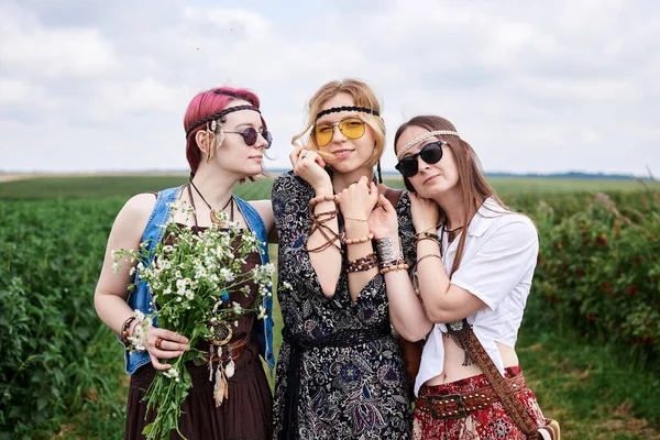 Üç genç hippi kadın, boho tarzı kıyafetler giyiyorlar, yeşil frenk üzümü tarlasında duruyorlar, yazın papatya buketi tutuyorlar. Eko turizm konsepti. Kırsal kesimde seyahat eden arkadaşlar.
