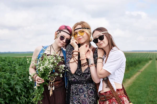 Üç genç hippi kadın, boho tarzı kıyafetler giyiyorlar, yeşil frenk üzümü tarlasında duruyorlar, yazın papatya buketi tutuyorlar. Eko turizm konsepti. Kırsal kesimde seyahat eden arkadaşlar.