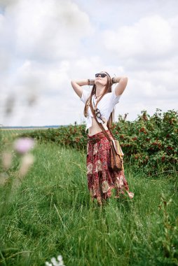 Genç esmer hippi kadın, boho tarzı kıyafetler ve güneş gözlüğü takıyor, yeşil frenk üzümü tarlasında duruyor, ellerini uzatıyor. Doğal arka planda bir kadın portresi. Çevre turizmi kavramı.