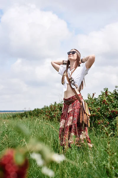 Young brunette hippie woman, wearing boho style clothes and sunglasses, standing on green currant field, stretching hands. Female portrait on natural background. Eco tourism concept.