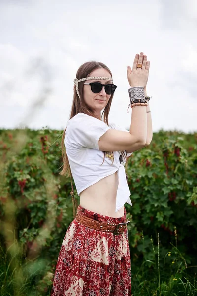 Young brunette hippie woman, wearing boho style clothes and sunglasses, standing on green currant field, with hands up, in dance pose. Female portrait on natural background. Eco tourism concept.