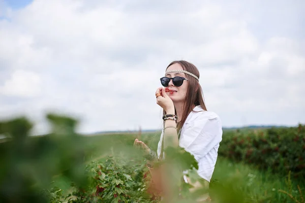 年轻的黑发嬉皮士女人 身穿布荷风格的衣服 戴着太阳镜 拿起并吃着绿色田野上的红醋栗浆果 生态旅游概念 — 图库照片
