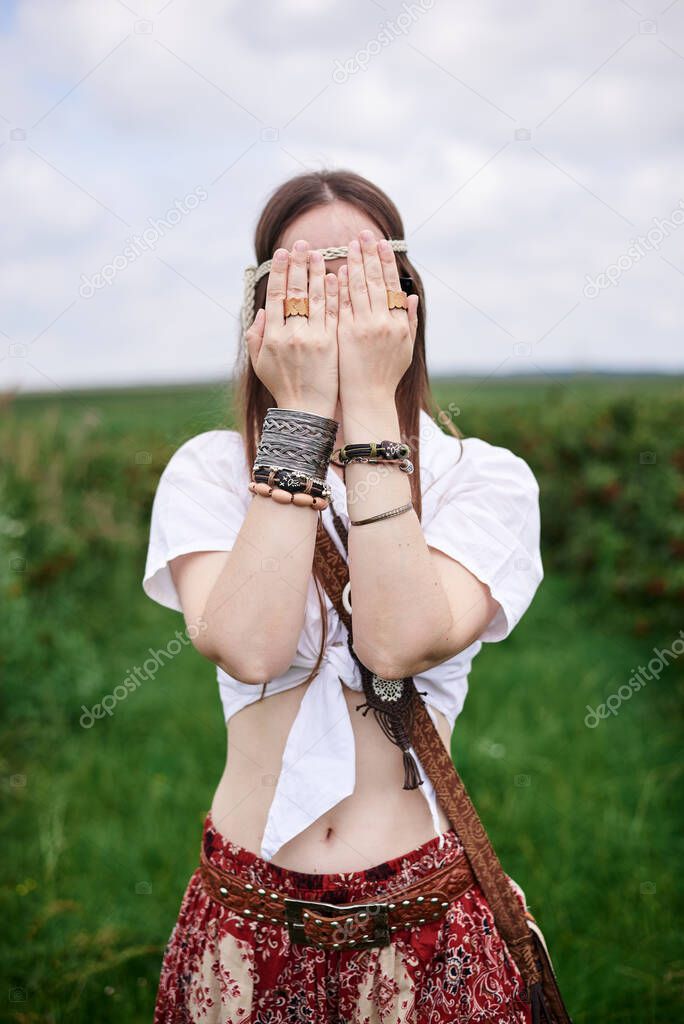 Young brunette hippie woman, wearing boho style clothes and sunglasses, standing on green currant field, covering face with hands with bracelets. Female portrait on natural background. Eco tourism.