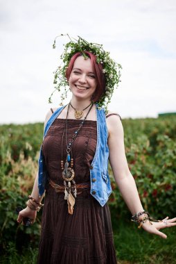 Kısa kızıl saçlı, boho tarzı kıyafetler ve çiçek çelengi giyen, yeşil frenk üzümü tarlasında duran, gülümseyen, gülen genç hippi kadın. Doğal arka planda bir kadın portresi. Çevre turizmi kavramı.