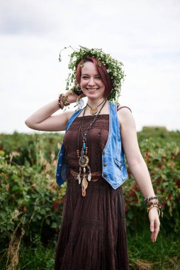 Kısa kızıl saçlı, boho tarzı kıyafetler ve çiçek çelengi giyen, yeşil frenk üzümü tarlasında duran, gülümseyen, gülen genç hippi kadın. Doğal arka planda bir kadın portresi. Çevre turizmi kavramı.