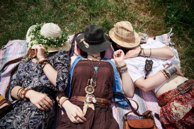 Üç hippi kadın, renkli boho tarzı kıyafetler ve hasır şapkalar giyiyorlar güneşli yaz gününde yeşil çimlerde uzanıp dinleniyorlar. Kırsal kesimde çevre turizmi. Birlikte seyahat eden arkadaşlar..