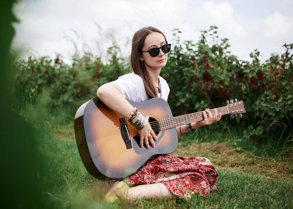 Young brunette hippie woman, wearing boho style clothes, sitting on green grass, holding guitar. Indie musician relaxing on green currant field on sunny summer day. Eco tourism concept.