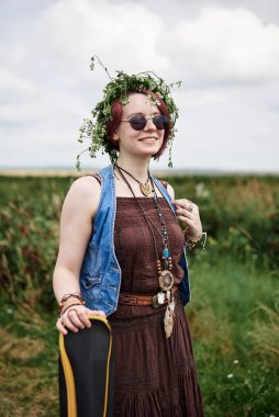 Kısa kızıl saçlı hippi kadın, boho tarzı kıyafetler, güneş gözlüğü ve çiçek çelengi takıyor, yeşil tarlada duruyor, gitar tutuyor. Kırsalda bağımsız müzisyen. Çevre turizmi kavramı.