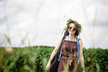 Kısa kızıl saçlı hippi kadın, boho tarzı kıyafetler, güneş gözlüğü ve çiçek çelengi takıyor, yeşil tarlada duruyor, gitar tutuyor. Kırsalda bağımsız müzisyen. Çevre turizmi kavramı.