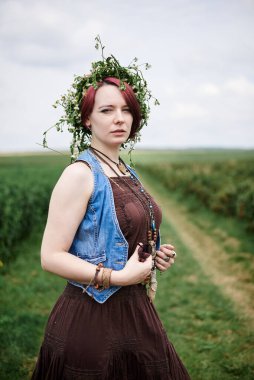 Kısa kızıl saçlı, boho tarzı kıyafetler ve çiçek çelengi giyen, yeşil frenk üzümü tarlasında duran, resim için poz veren genç hippi kadın. Doğal arka planda bir kadın portresi. Çevre turizmi kavramı.