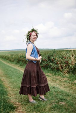 Kısa kızıl saçlı, boho tarzı kıyafetler ve çiçek çelengi giyen, yeşil frenk üzümü tarlasında duran, resim için poz veren genç hippi kadın. Doğal arka planda bir kadın portresi. Çevre turizmi kavramı.