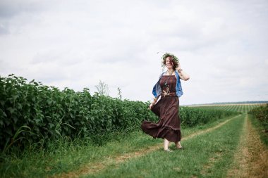 Kısa kızıl saçlı hippi kadın, boho tarzı kıyafetler ve çiçek çelengi giyiyor, yeşil alanda koşuyor, gülümsüyor, gülüyor. Doğal arka planda bir kadın portresi. Çevre turizmi kavramı.