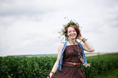 Kısa kızıl saçlı hippi kadın, boho tarzı kıyafetler ve çiçek çelengi giyiyor, yeşil alanda koşuyor, gülümsüyor, gülüyor. Doğal arka planda bir kadın portresi. Çevre turizmi kavramı.