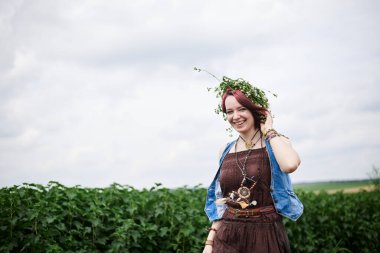 Kısa kızıl saçlı hippi kadın, boho tarzı kıyafetler ve çiçek çelengi giyiyor, yeşil alanda koşuyor, gülümsüyor, gülüyor. Doğal arka planda bir kadın portresi. Çevre turizmi kavramı.