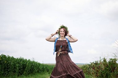 Kısa kızıl saçlı hippi kadın, boho tarzı kıyafetler ve çiçek çelengi giyiyor, yeşil alanda koşuyor, gülümsüyor, gülüyor. Doğal arka planda bir kadın portresi. Çevre turizmi kavramı.