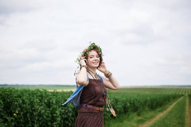 Kısa kızıl saçlı hippi kadın, boho tarzı kıyafetler ve çiçek çelengi giyiyor, yeşil alanda koşuyor, gülümsüyor, gülüyor. Doğal arka planda bir kadın portresi. Çevre turizmi kavramı.