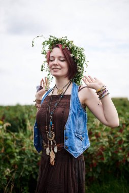 Kısa kızıl saçlı, boho tarzı kıyafetler ve çiçek çelengi giyen, yeşil frenk üzümü tarlasında duran, gülümseyen, gülen genç hippi kadın. Doğal arka planda bir kadın portresi. Çevre turizmi kavramı.